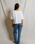 A person with long dark hair, wearing the Perfectwhitetee French Terry SS Loose Sweatshirt in white and blue jeans, stands facing away from the camera against a neutral draped fabric background.