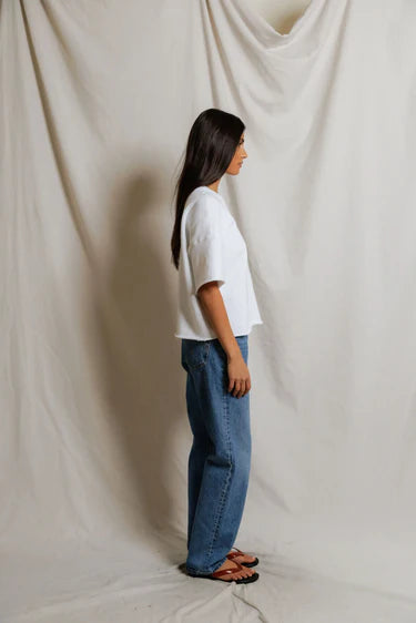 A woman stands in profile against a light beige fabric backdrop, wearing the Perfectwhitetee French Terry SS Loose Sweatshirt, blue jeans, and black sandals. Her straight dark hair is worn down.