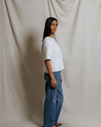 A woman with long dark hair stands sideways, looking at the camera. She wears the Perfectwhitetee Ezra SS Terry Sweatshirt in white, with blue jeans and brown sandals against a cream-colored fabric backdrop.