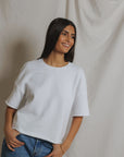 A woman with long dark hair, wearing the Perfectwhitetee Ezra SS Terry Sweatshirt in white and blue jeans, stands smiling with one hand in her pocket against a neutral fabric backdrop.