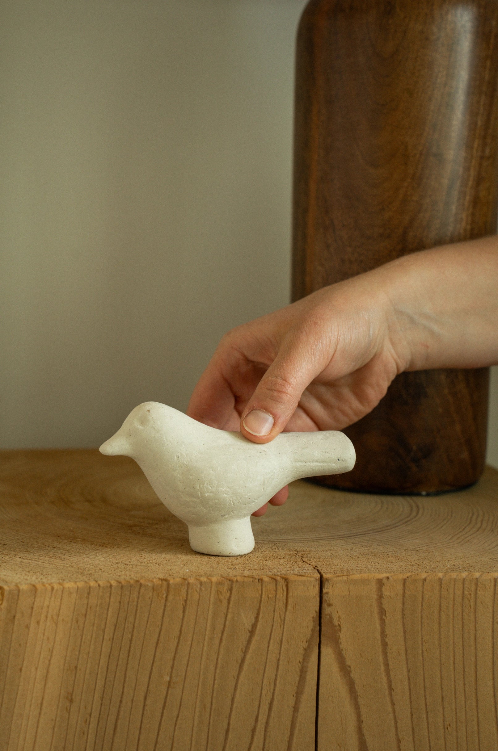 A hand gently places the Indaba Paper Mache Bird White accent decor on a wooden surface, with a tall wooden vase in the background.