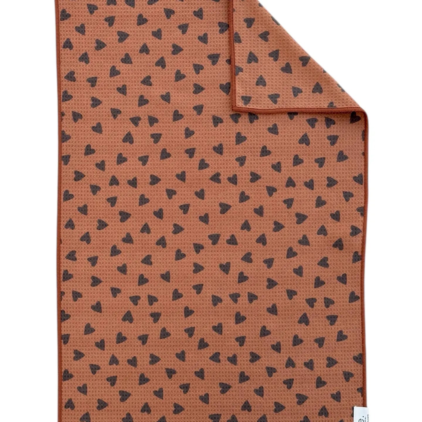 The Faire Double Sided Hand Towel features a brown rectangular design with small black hearts scattered across both sides; one corner is folded to show the reversible pattern.