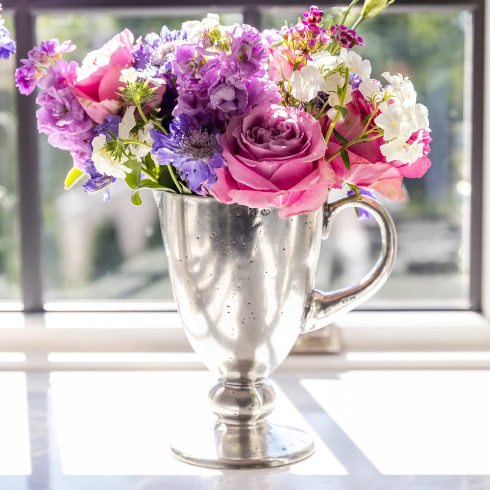 The Veneto Pitcher by Faire holds a vibrant bouquet of pink roses, purple, and white flowers, displayed on a sunlit windowsill with greenery softly blurred in the background.