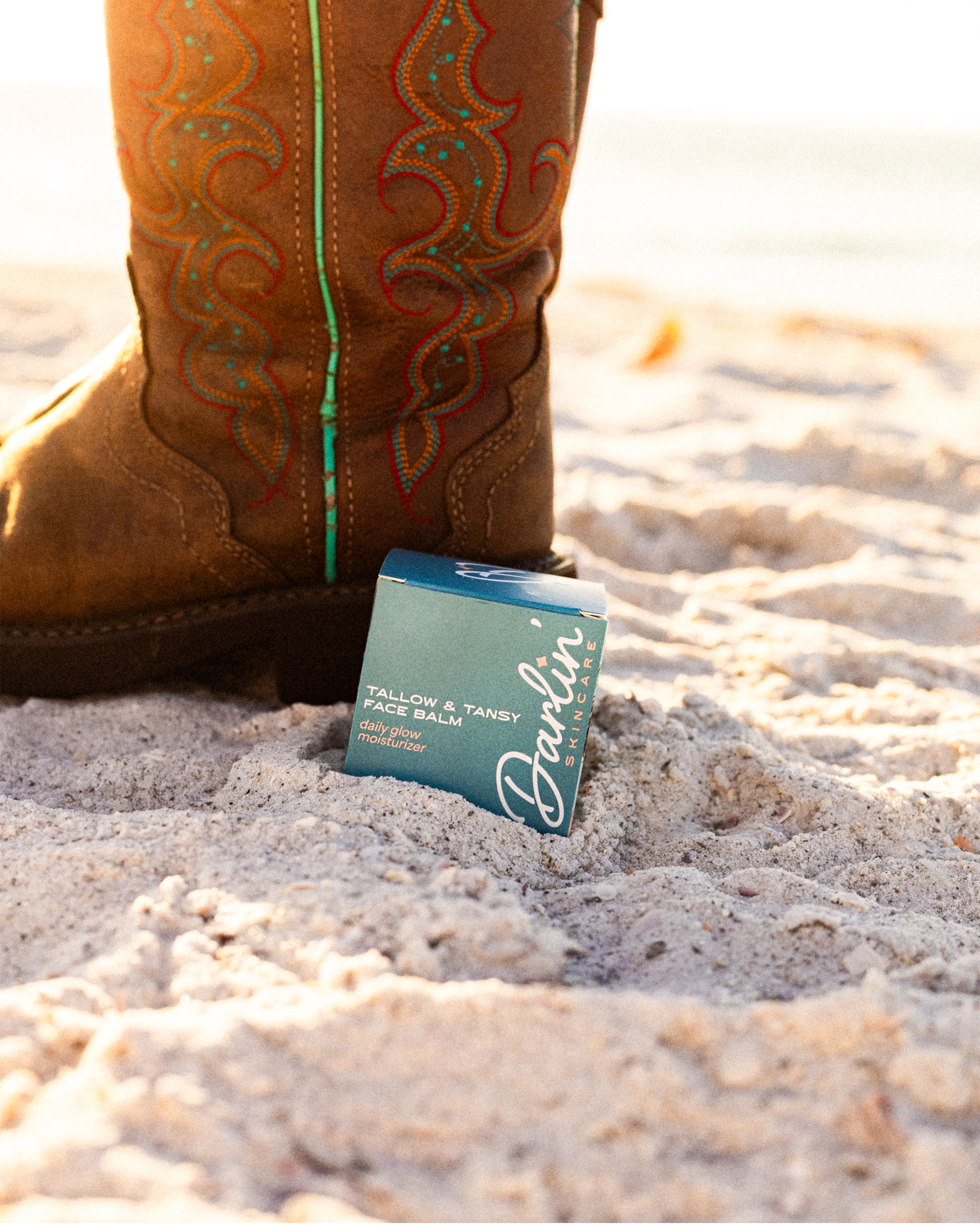 A close-up of a person in a brown cowboy boot with colorful stitching stands on sandy ground beside a blue Faire box labeled “Tallow Face Balm Moisturizer,” featuring Blue Tansy and grass-fed tallow for anti-aging skincare.