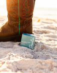 A close-up of a person in a brown cowboy boot with colorful stitching stands on sandy ground beside a blue Faire box labeled “Tallow Face Balm Moisturizer,” featuring Blue Tansy and grass-fed tallow for anti-aging skincare.