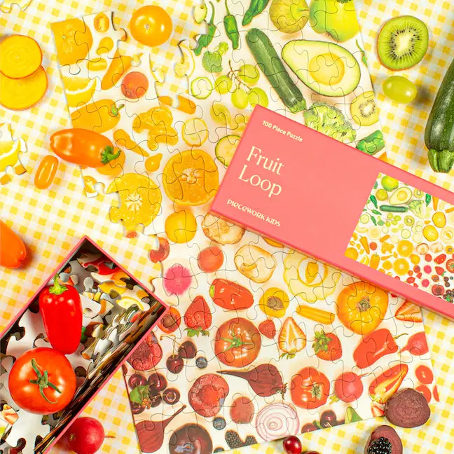 The Faire Kids 100 Piece Puzzle features a vibrant fruit and veggie theme on a yellow gingham background, surrounded by real tomatoes, peppers, and kiwi. Crafted on high-quality art paper, the puzzle box is displayed invitingly on top.