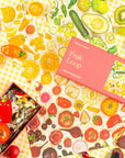 The Faire Kids 100 Piece Puzzle features a vibrant fruit and veggie theme on a yellow gingham background, surrounded by real tomatoes, peppers, and kiwi. Crafted on high-quality art paper, the puzzle box is displayed invitingly on top.