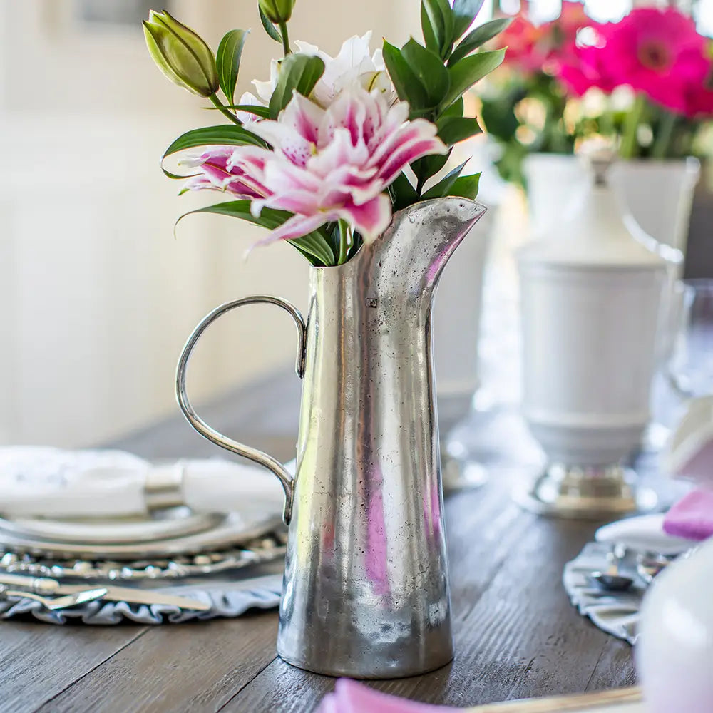A Vintage Tall Tapered Pitcher by Faire, filled with pink and white flowers, sits on a set dining table featuring white plates, napkins, and additional floral centerpieces in the background.