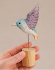 Purple hand-painted carved hummingbird figurine on wood base by Faire, shown held in hand for scale.