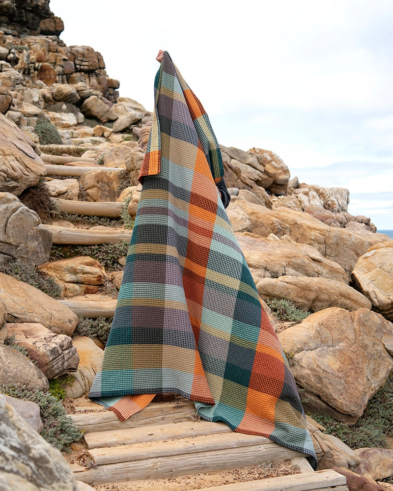 Outdoors on rocky steps, a person is mostly covered by the Vrou-Vrou Blanket from Faire, its green, orange, brown, and cream checks blending seamlessly into the rugged natural surroundings.