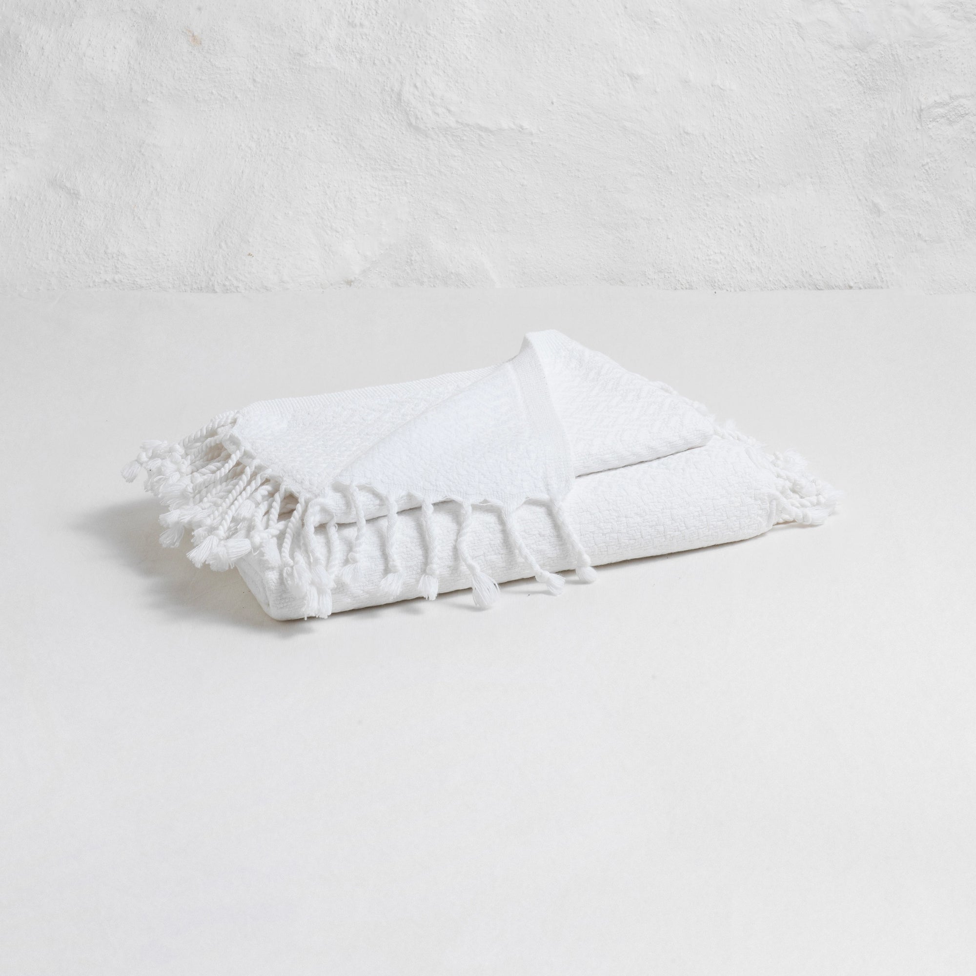 The Faire Spa Hand Towel, crafted from Turkish cotton with fringed edges, is neatly folded on a white surface in front of a textured white wall.