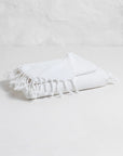 The Faire Spa Hand Towel, crafted from Turkish cotton with fringed edges, is neatly folded on a white surface in front of a textured white wall.