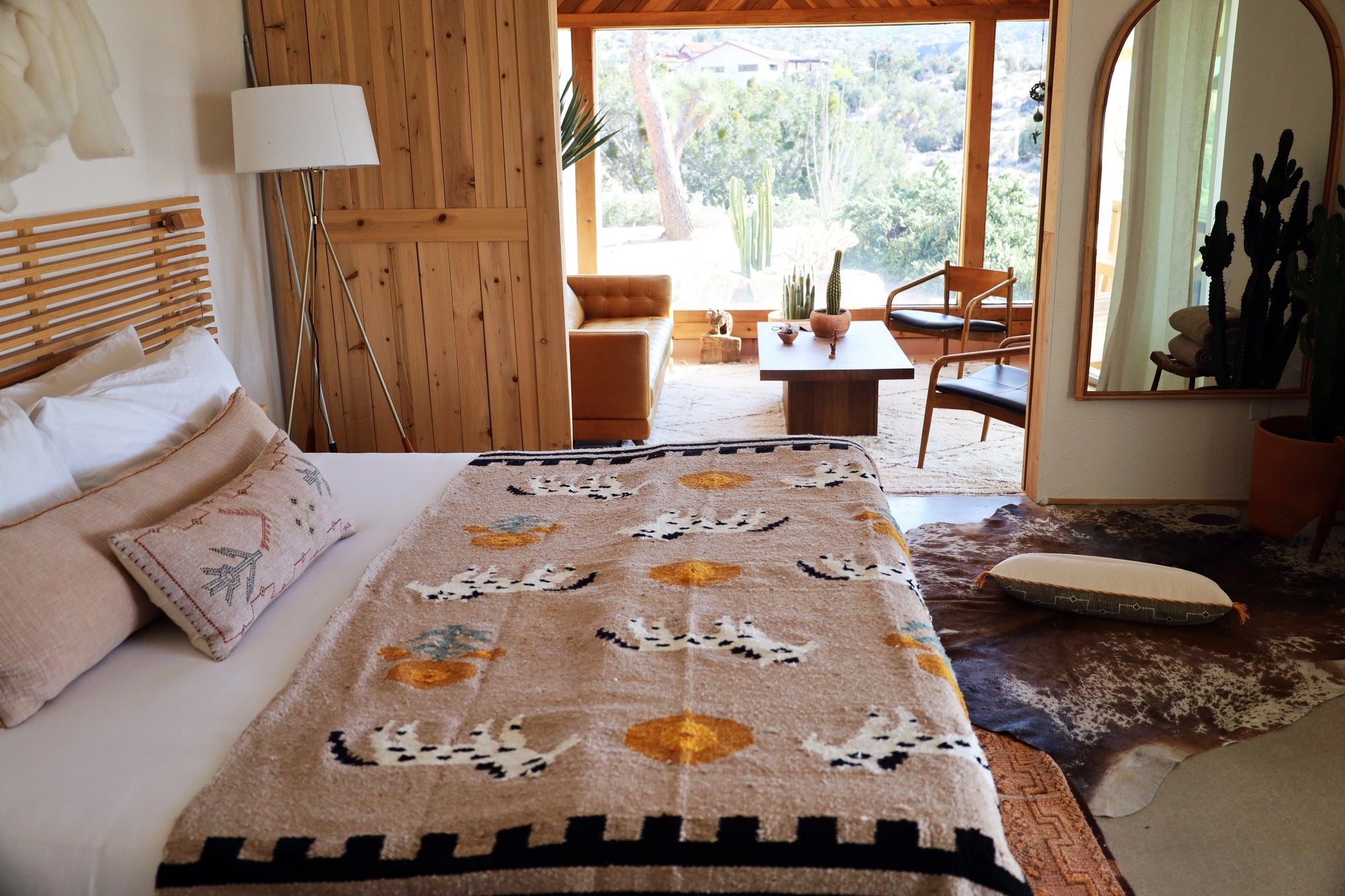 A cozy bedroom showcases the TT Handwoven Blanket Poppies n Ponies by Faire atop a bed with beige pillows, wooden headboard, and lamp. Nearby, armchairs and plants sit by large windows overlooking lush greenery.