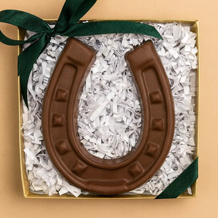 The Faire Chocolate Horseshoe, crafted in the USA, rests on white shredded paper in a gold gift box finished with a dark green ribbon—a delicious symbol of good luck.