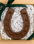 The Faire Chocolate Horseshoe, crafted in the USA, rests on white shredded paper in a gold gift box finished with a dark green ribbon—a delicious symbol of good luck.