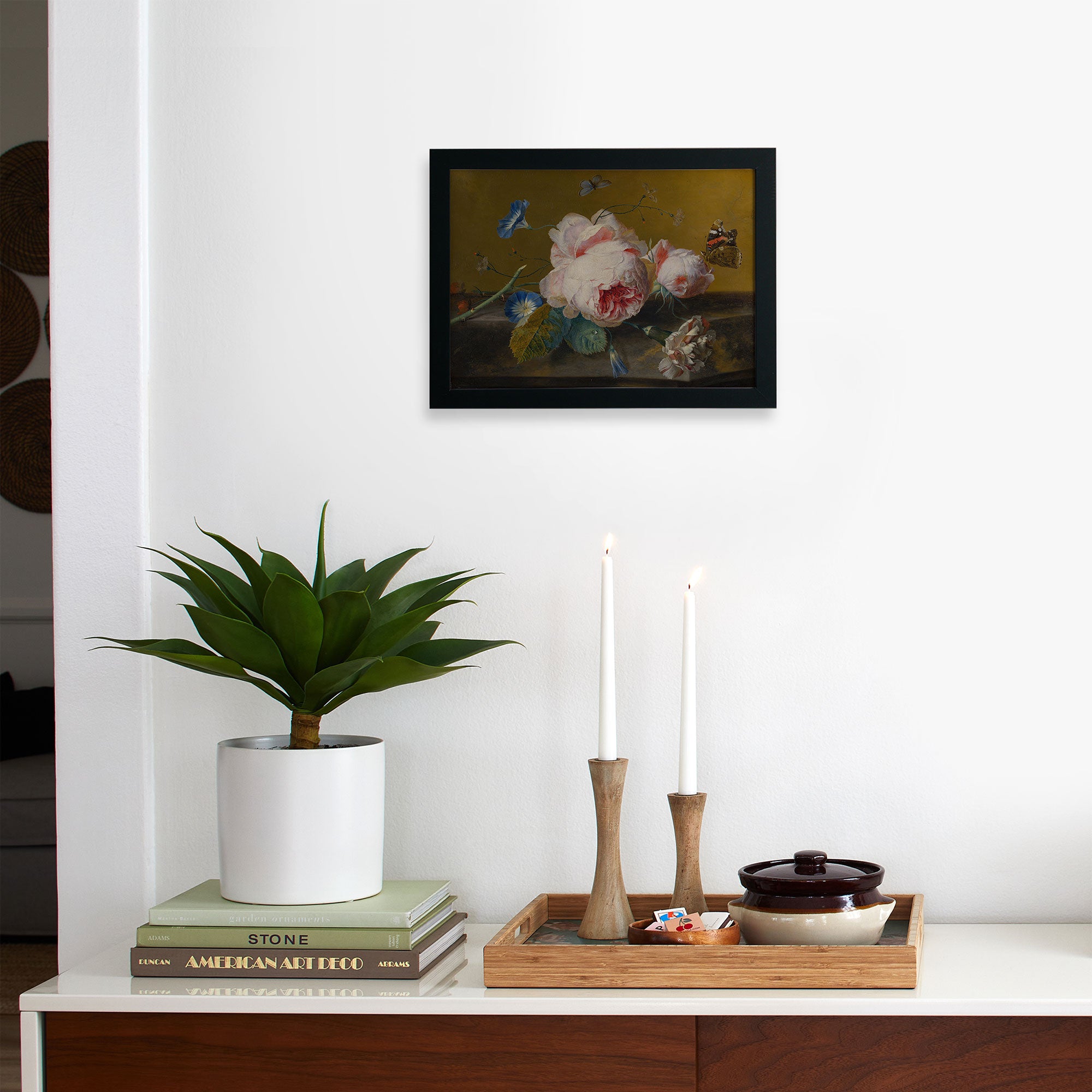 The Faire Van Huysum Flower Still Framed art hangs on a white wall above a shelf with a potted plant, two lit wooden-cased candles, stacked books, and a tray that holds a ceramic dish and small bowls.