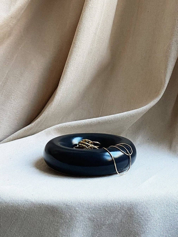 The Faire Gyrus Pocket Tray, a glossy black concrete piece with a cork base, holds rings and a gold necklace atop cream textured fabric with soft folds, offering both style and surface protection.