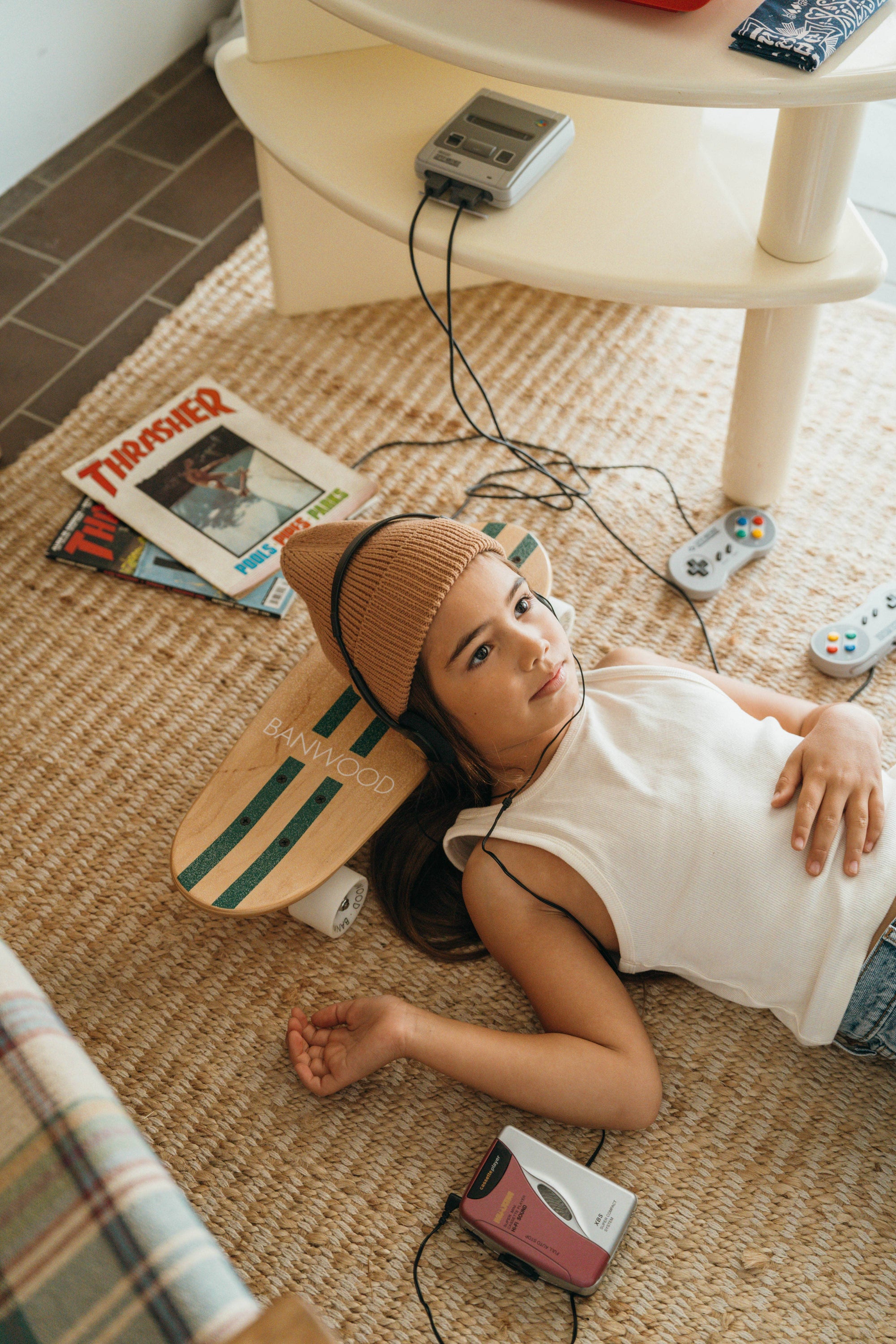 A girl in a beanie and tank top relaxes on a woven rug with headphones, resting her head on the BANWOOD SKATEBOARD - GREEN by Faire. Vintage video game controllers, a cassette player, and skateboard magazines are nearby.