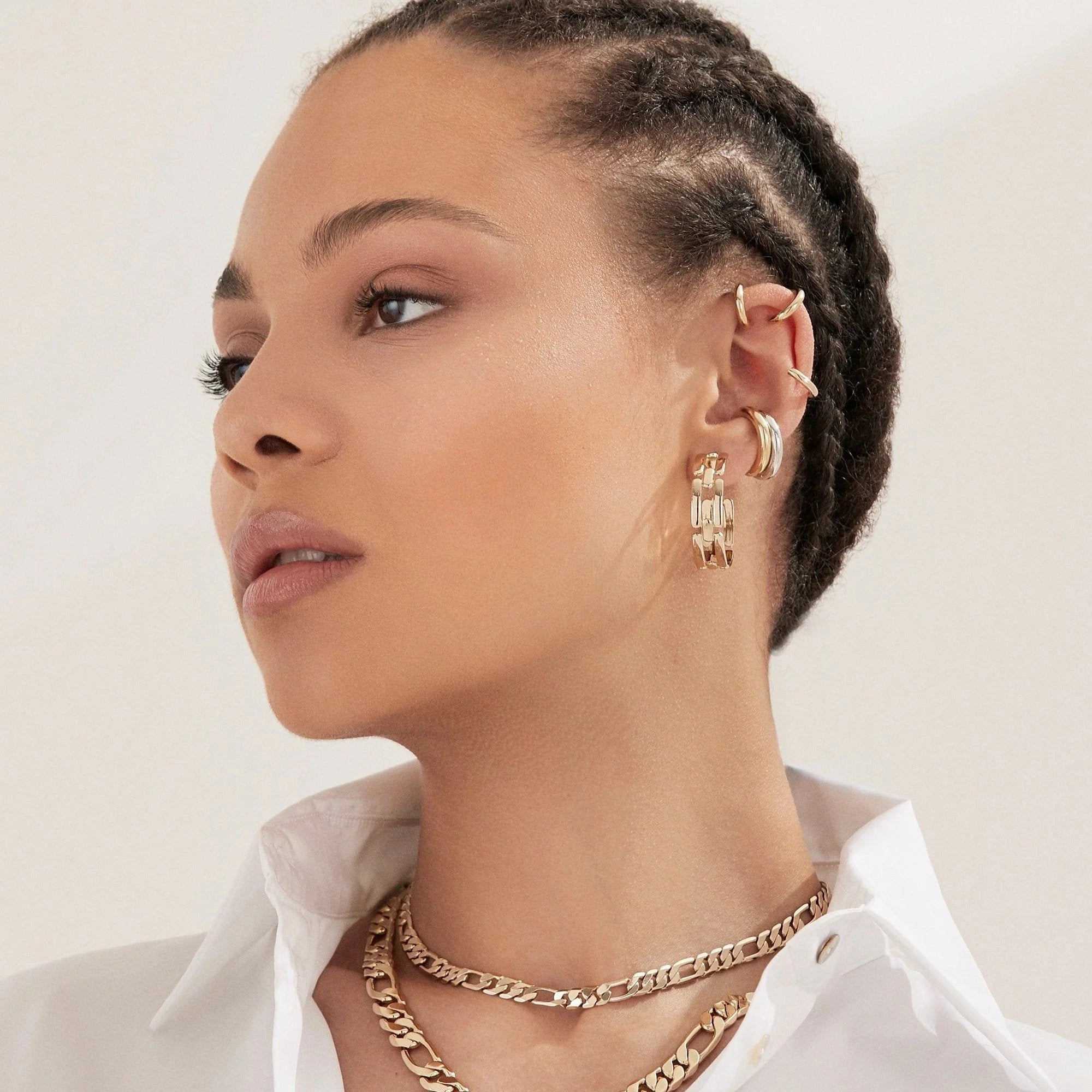 A woman with braided hair wears several gold earrings—including Faire’s Lisa Ear Cuff in 10K gold on her mid-ear cartilage—plus layered gold necklaces and a white collared shirt, glancing to the side against a neutral background.