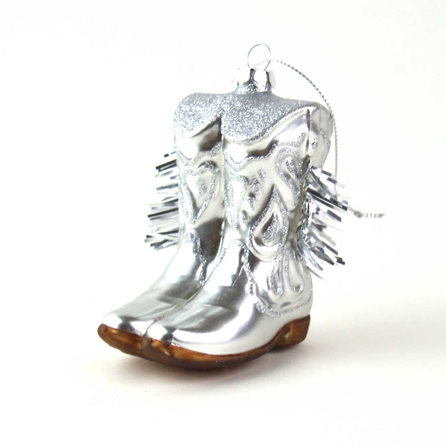 The Faire Cowgirl Boot Ornament features shiny silver boots with decorative swirls, spiky accents, brown soles, and a hanging loop. This glossy Western décor adds festive charm to any tree.