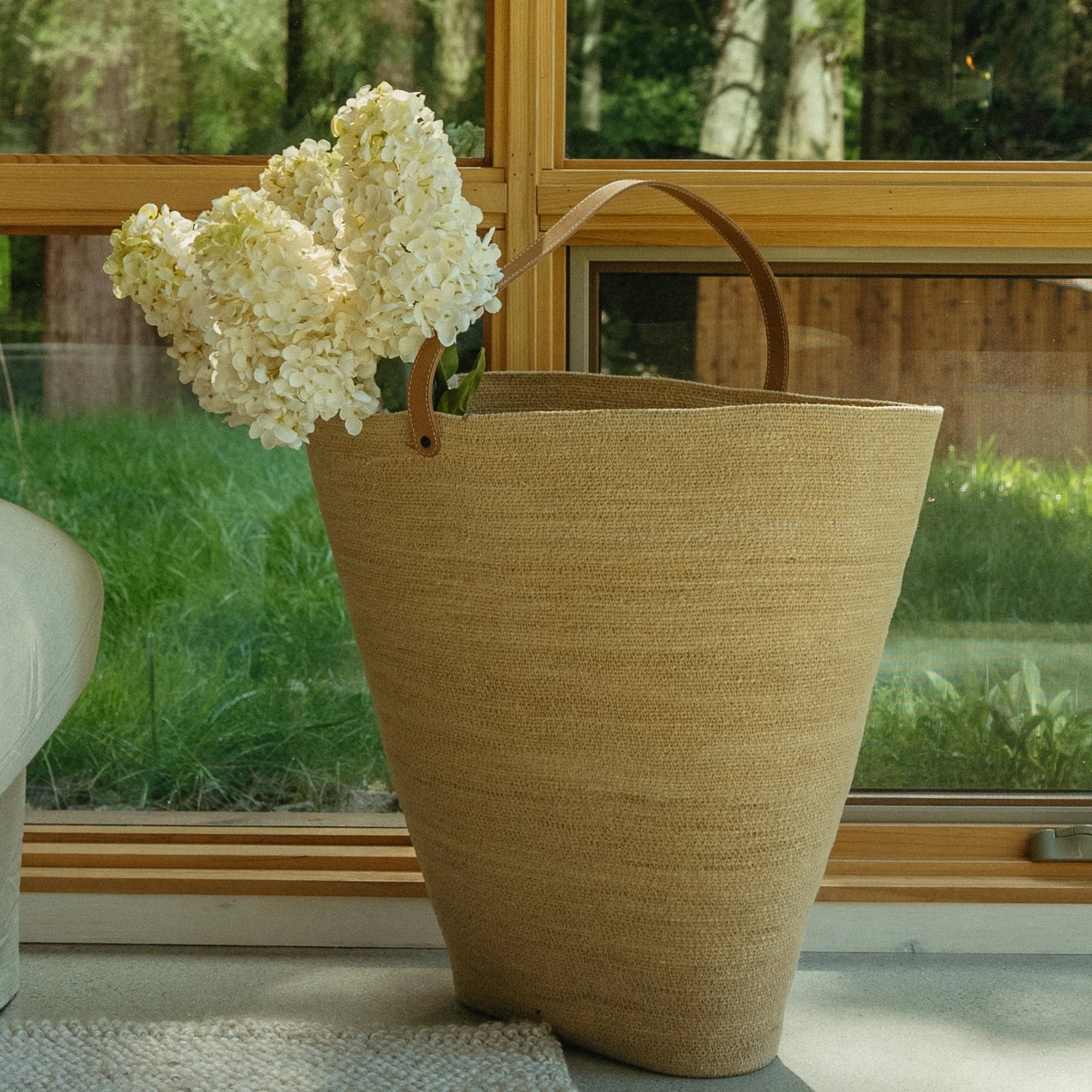 An Indaba Hydrangea Paniculata Stem is arranged in a large woven basket with a leather handle, set indoors near a window overlooking sunlit green trees.