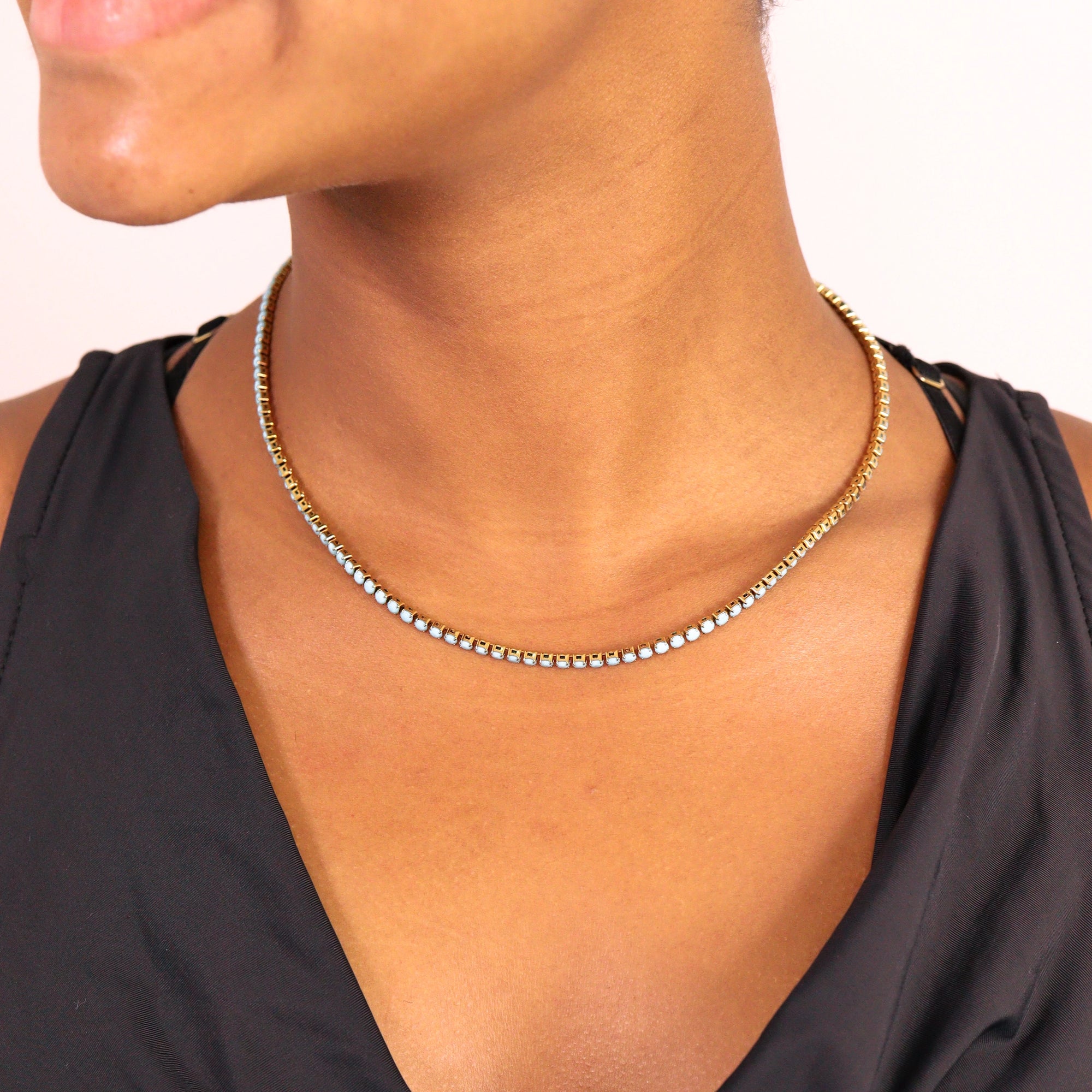 A woman wearing a black v-neck top and the Faire Turquoise Tennis Necklace, featuring small white beads and made from hypoallergenic 316L stainless steel, shown from chin to chest.