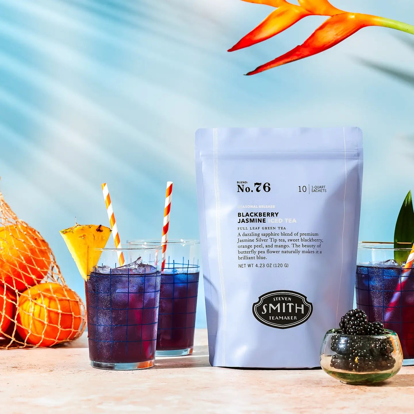 A package of Smith Tea by Faire sits on a table with three glasses of refreshing iced tea. Each drink is served with striped straws, garnished with blackberries and ice, against a backdrop of fresh oranges and hibiscus flowers.