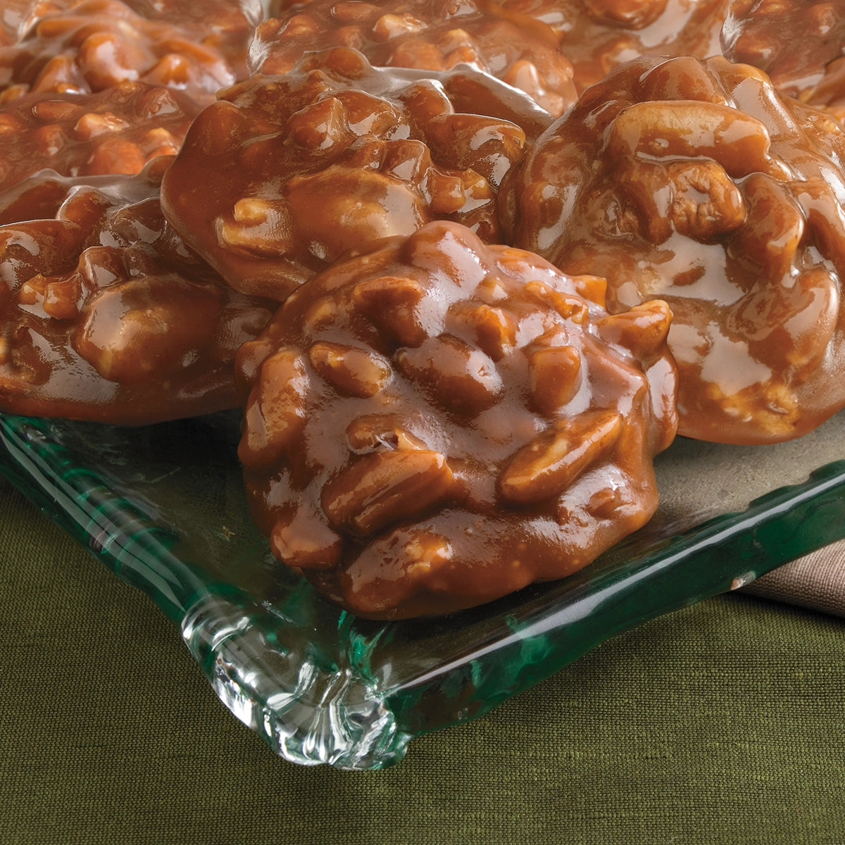A close-up of Faire’s LC Texas Chewie Pecan Pralines 6oz, glossy and rich brown, stacked on a clear glass plate atop green fabric. Each all-natural candy is filled with whole pecans.