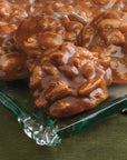 A close-up of Faire’s LC Texas Chewie Pecan Pralines 6oz, glossy and rich brown, stacked on a clear glass plate atop green fabric. Each all-natural candy is filled with whole pecans.