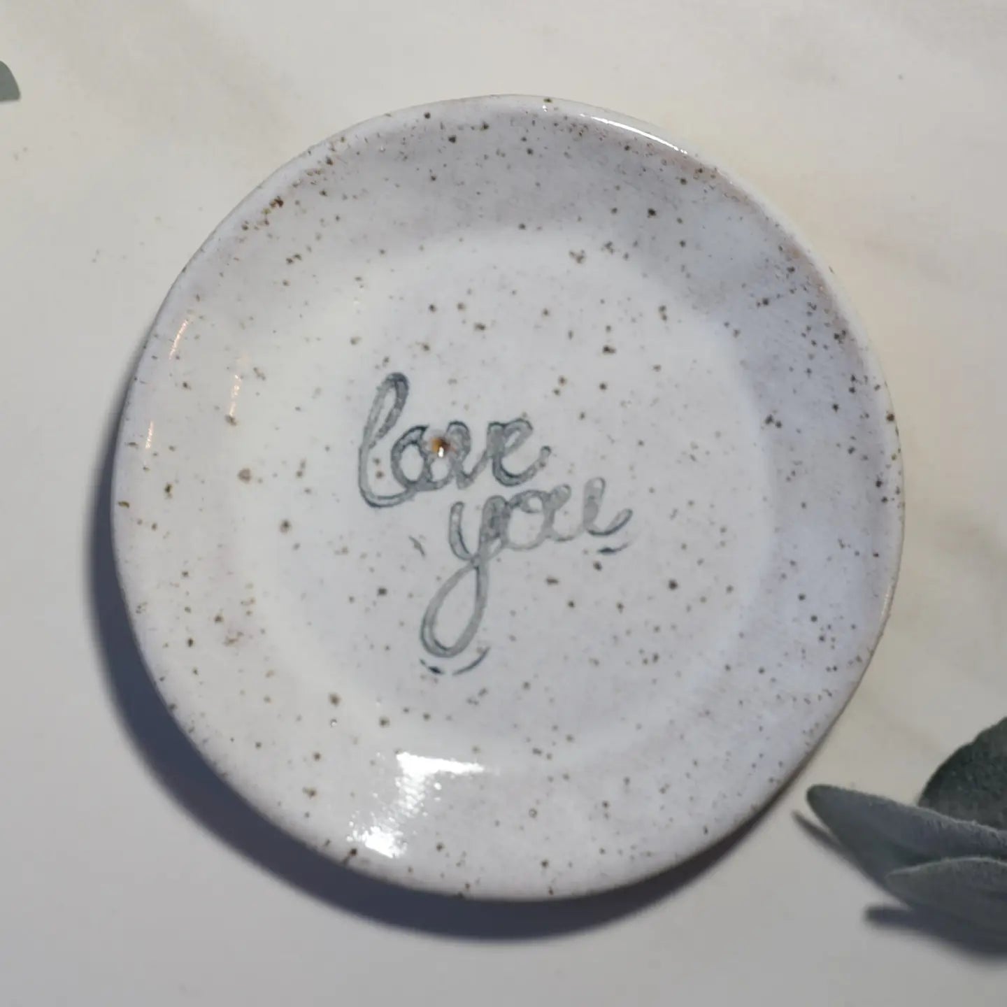 The Faire Mini Plate is a small, round, white ceramic dish with brown speckles and "love you" in cursive at the center. Ideal as a jewelry dish or for snacks. A greenish plant appears in the lower right corner of the image.