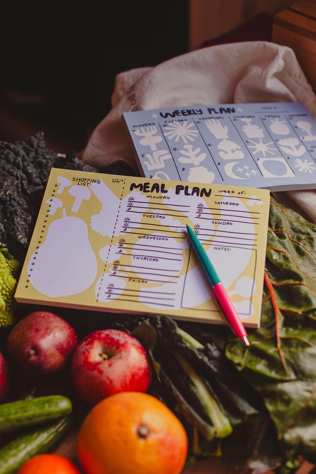 The Faire Meal Planner Notepad, made in the USA, is placed on leafy greens surrounded by apples, oranges, and cucumbers. Two pens rest on top.