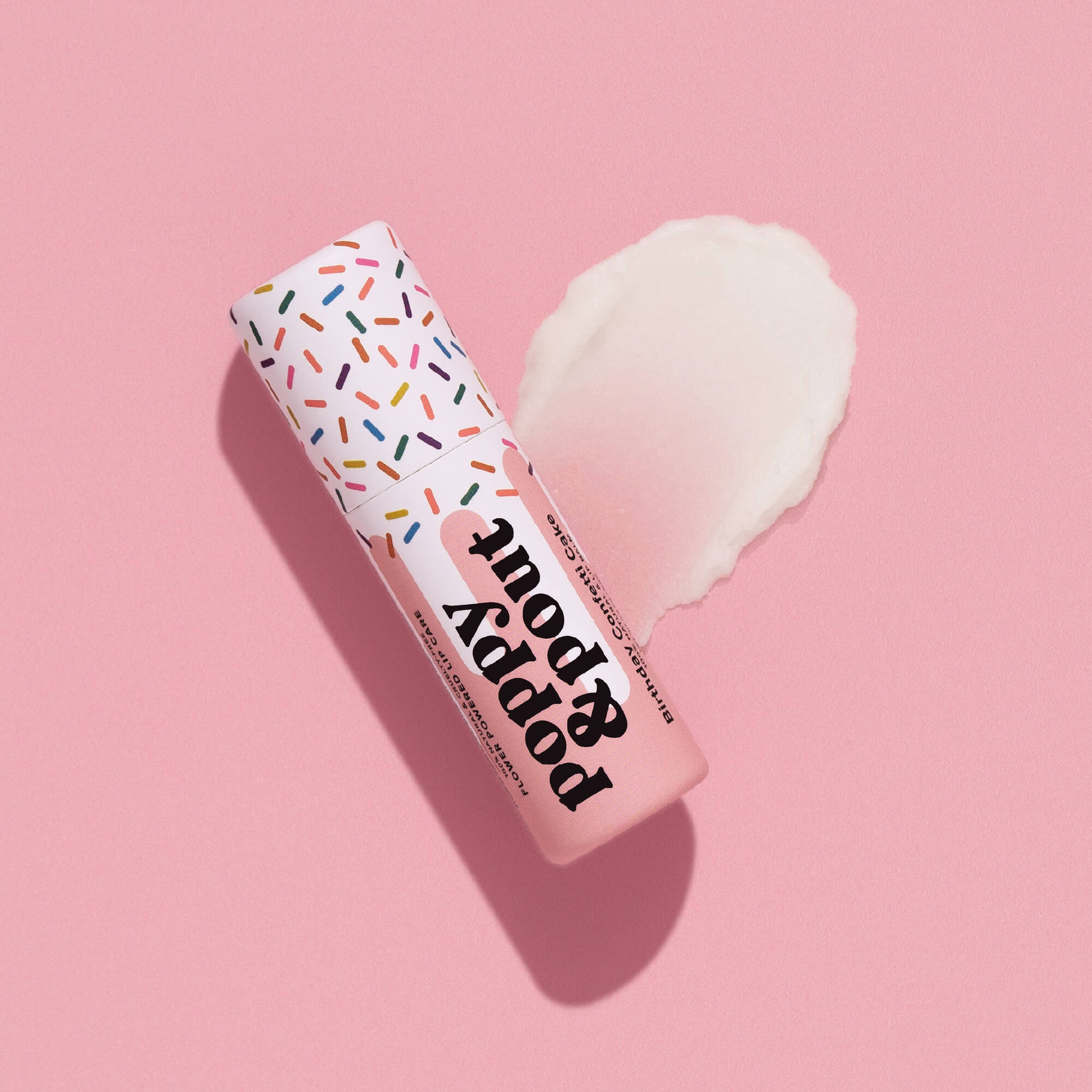 A pink tube of Faire's Poppy & Pout Lip Balm with a sprinkle-patterned cap rests on a pink background, next to a smear of white balm.