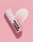 A pink tube of Faire's Poppy & Pout Lip Balm with a sprinkle-patterned cap rests on a pink background, next to a smear of white balm.
