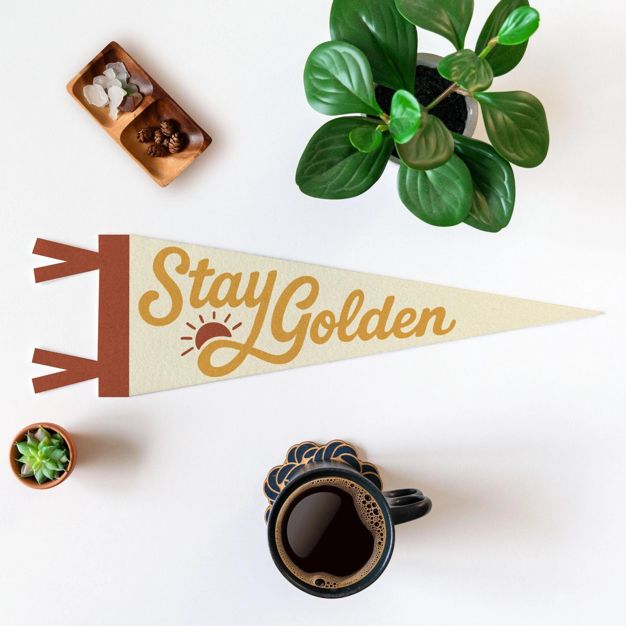 The Faire "Stay Golden" Felt Pennant, featuring a sun illustration in beige, brings handmade charm to any cozy space—ideal wall decor alongside coffee, plants, and crystals.