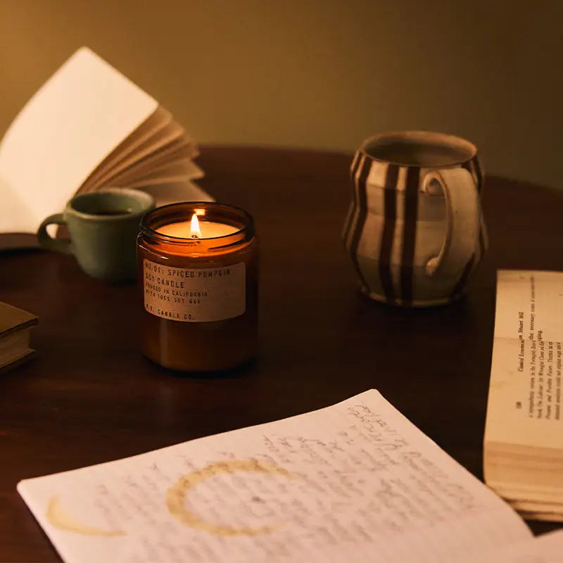 The PFC Candle by Faire glows warmly in a brown jar on a wooden table, surrounded by an open notebook, a striped ceramic mug, a green teacup, and handwritten notes—creating a cozy, inviting scene.