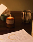 The PFC Candle by Faire glows warmly in a brown jar on a wooden table, surrounded by an open notebook, a striped ceramic mug, a green teacup, and handwritten notes—creating a cozy, inviting scene.