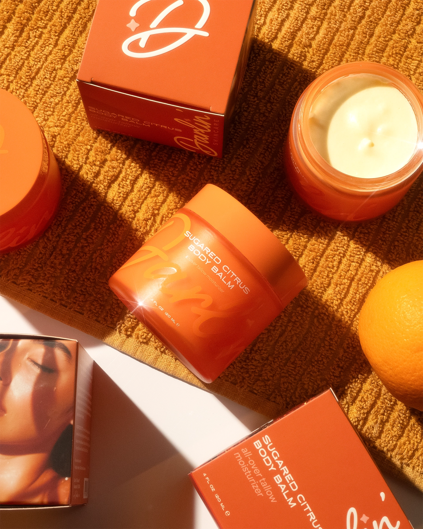 A jar of Faire's Tallow Body Butter Moisturizer in orange packaging rests on a textured orange towel, surrounded by its box, an open jar with balm, another box, and an orange fruit in warm natural light.