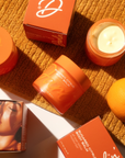 A jar of Faire's Tallow Body Butter Moisturizer in orange packaging rests on a textured orange towel, surrounded by its box, an open jar with balm, another box, and an orange fruit in warm natural light.