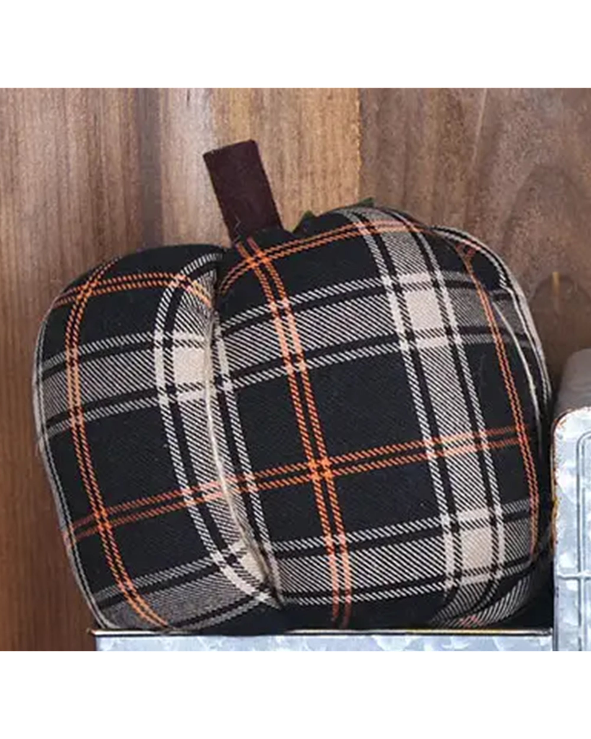 The Faire Pumpkin is a fabric ornament with black, beige, and orange plaid and a brown stem. Shown on a metal surface against wood, it makes the perfect farmhouse decor accent for autumn.
