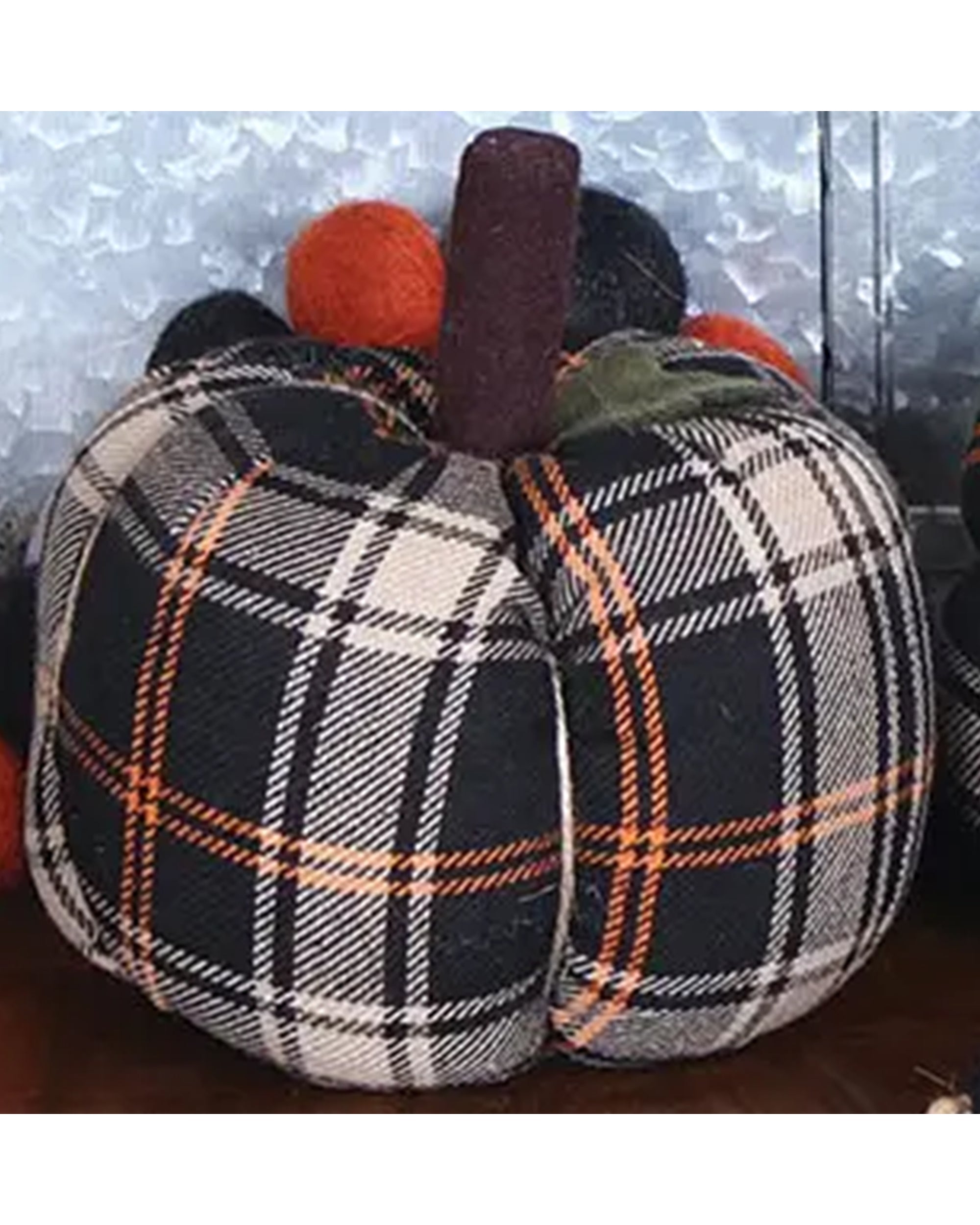 The Faire Pumpkin is a farmhouse decor piece with black, white, and orange plaid fabric, a brown stem, and round felt balls on top—a charming autumn ornament for your home against any metallic background.