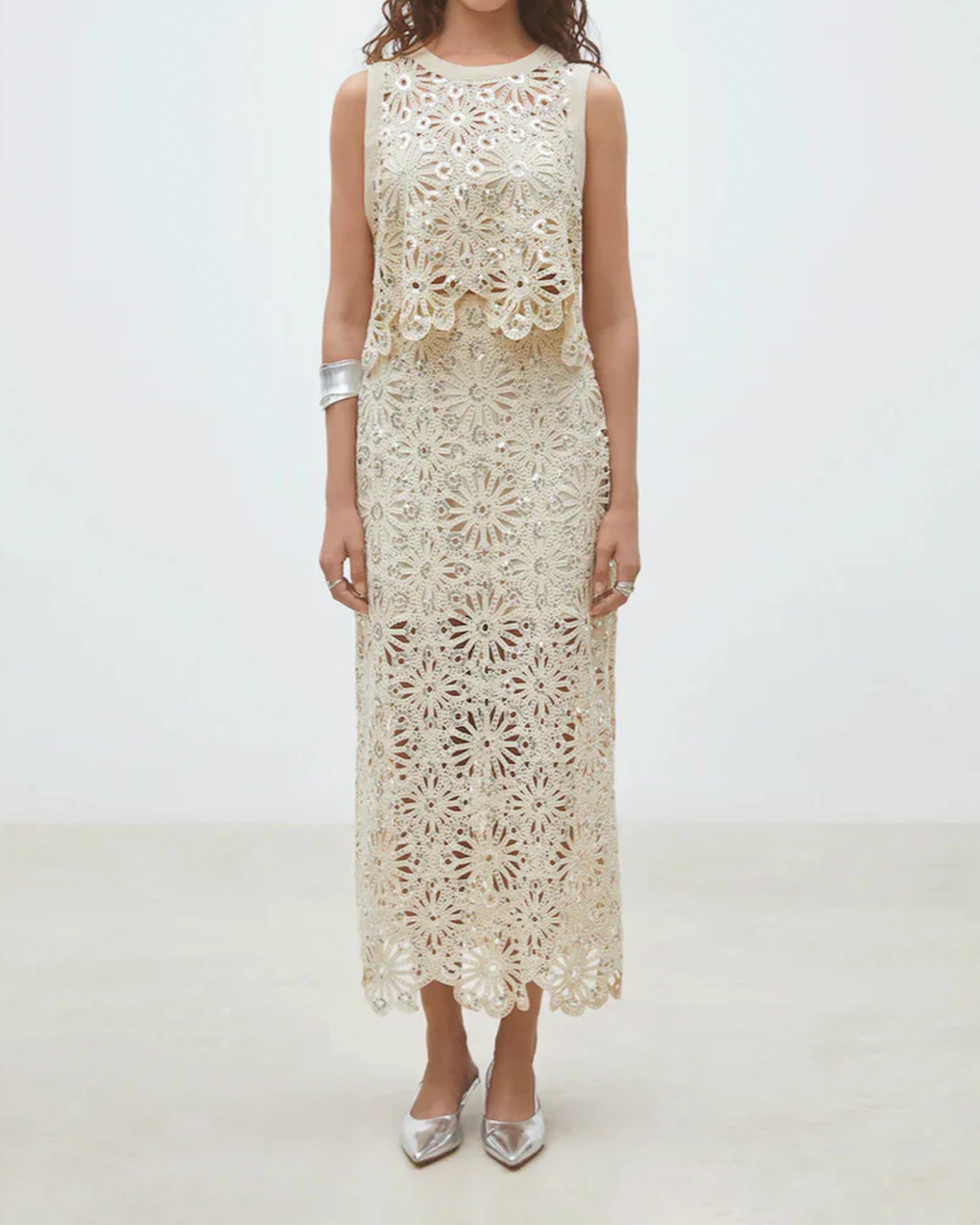 A woman wears the SUNCOO Fanny Skirt—a sleeveless, cream ankle-length skirt with lace detailing—paired with silver pointed-toe shoes and a chunky silver bracelet, posing against a plain light background.