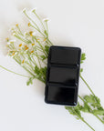 The Faire Travel Watercolor Paint Palette, a sleek black rectangular set, is displayed on white daisies and green leaves against a white background—an ideal gift for artists.