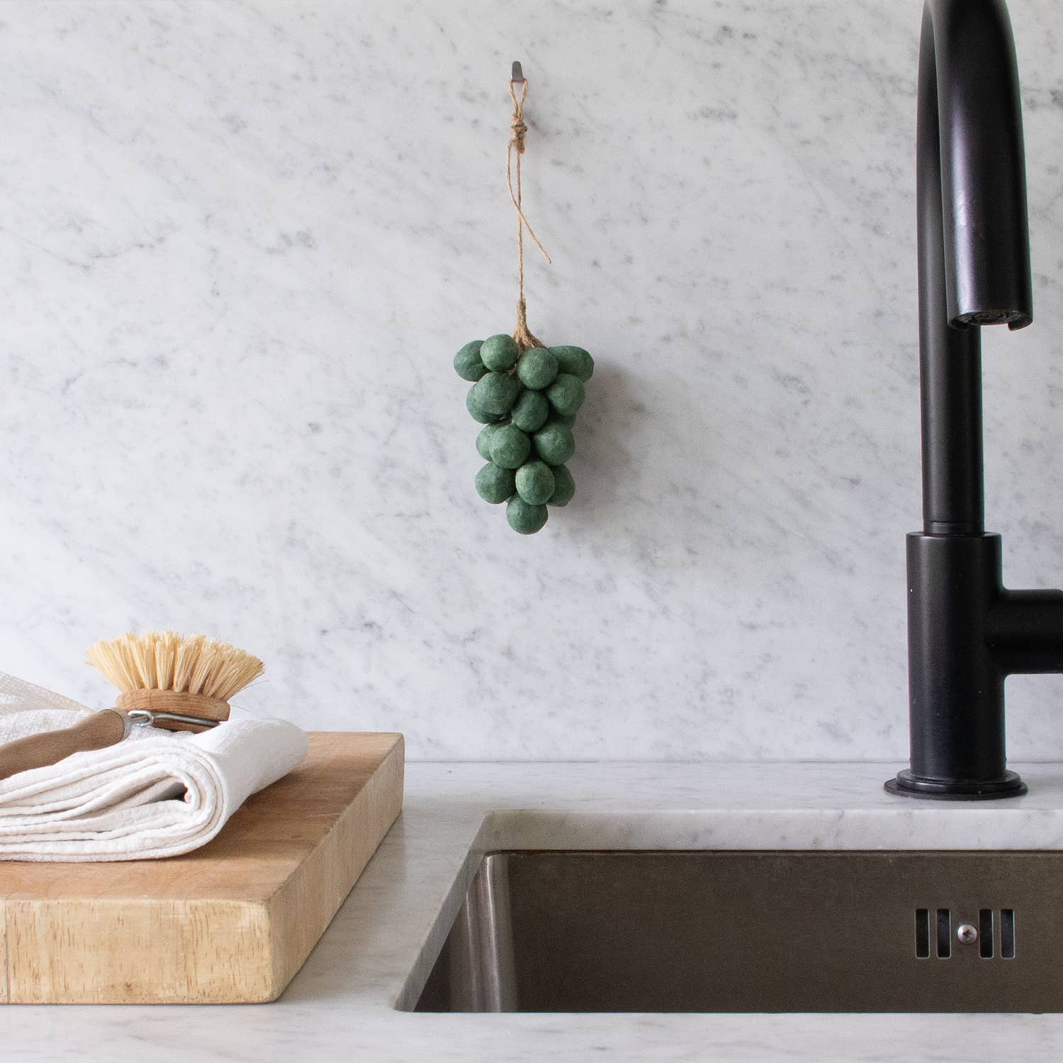 The Handcrafted Grapes Soap by Faire, shaped like a bunch of green grapes, hangs on a marble kitchen backsplash above a stainless steel sink, with vegan soap on a wooden cutting board and towels plus a dish brush nearby.