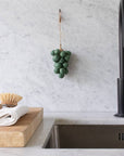 The Handcrafted Grapes Soap by Faire, shaped like a bunch of green grapes, hangs on a marble kitchen backsplash above a stainless steel sink, with vegan soap on a wooden cutting board and towels plus a dish brush nearby.