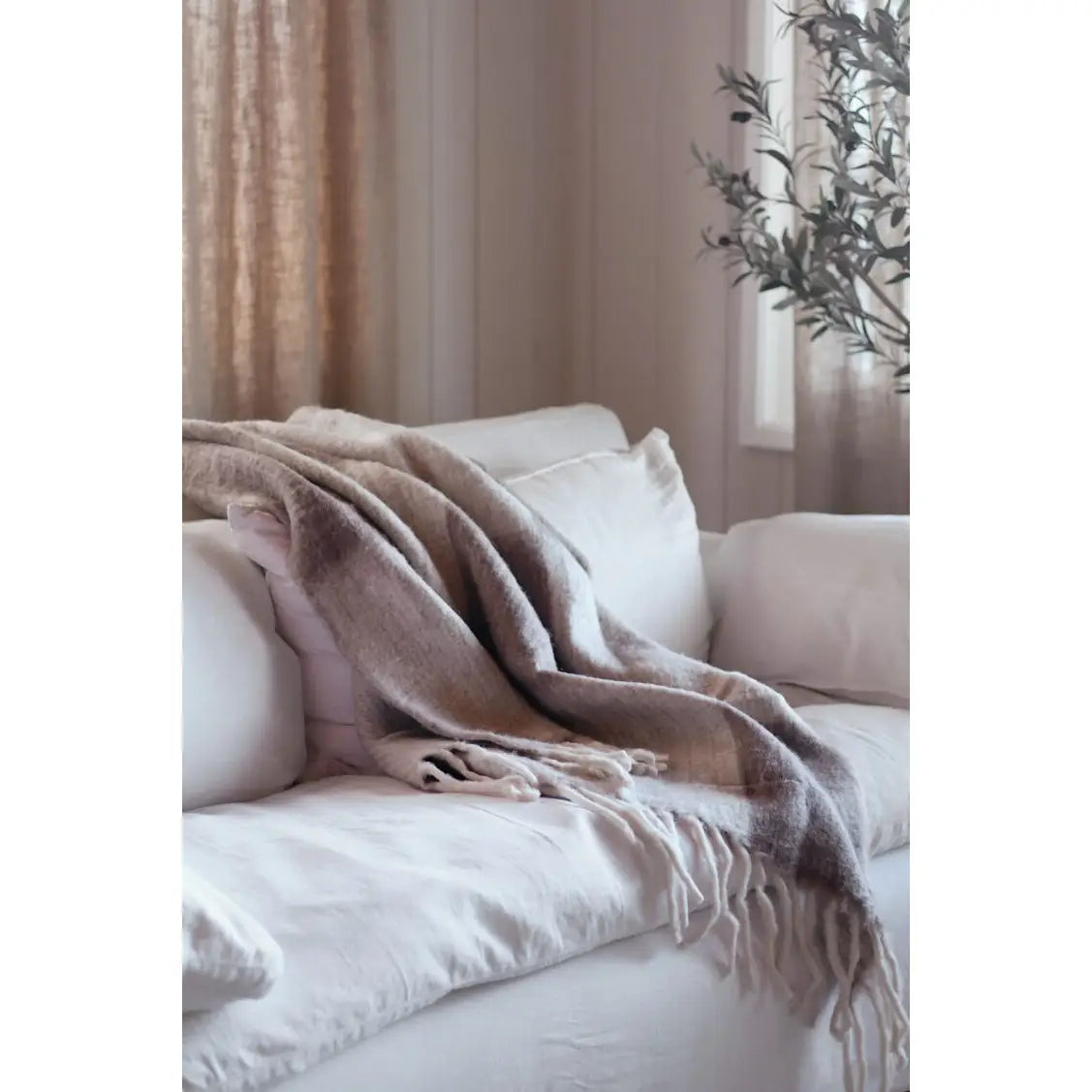 A cream sofa features the Indaba Checkerboard Throw in Taupe, its modern beige and brown pattern draped stylishly, set in a softly lit room with light walls and a leafy plant in the background.