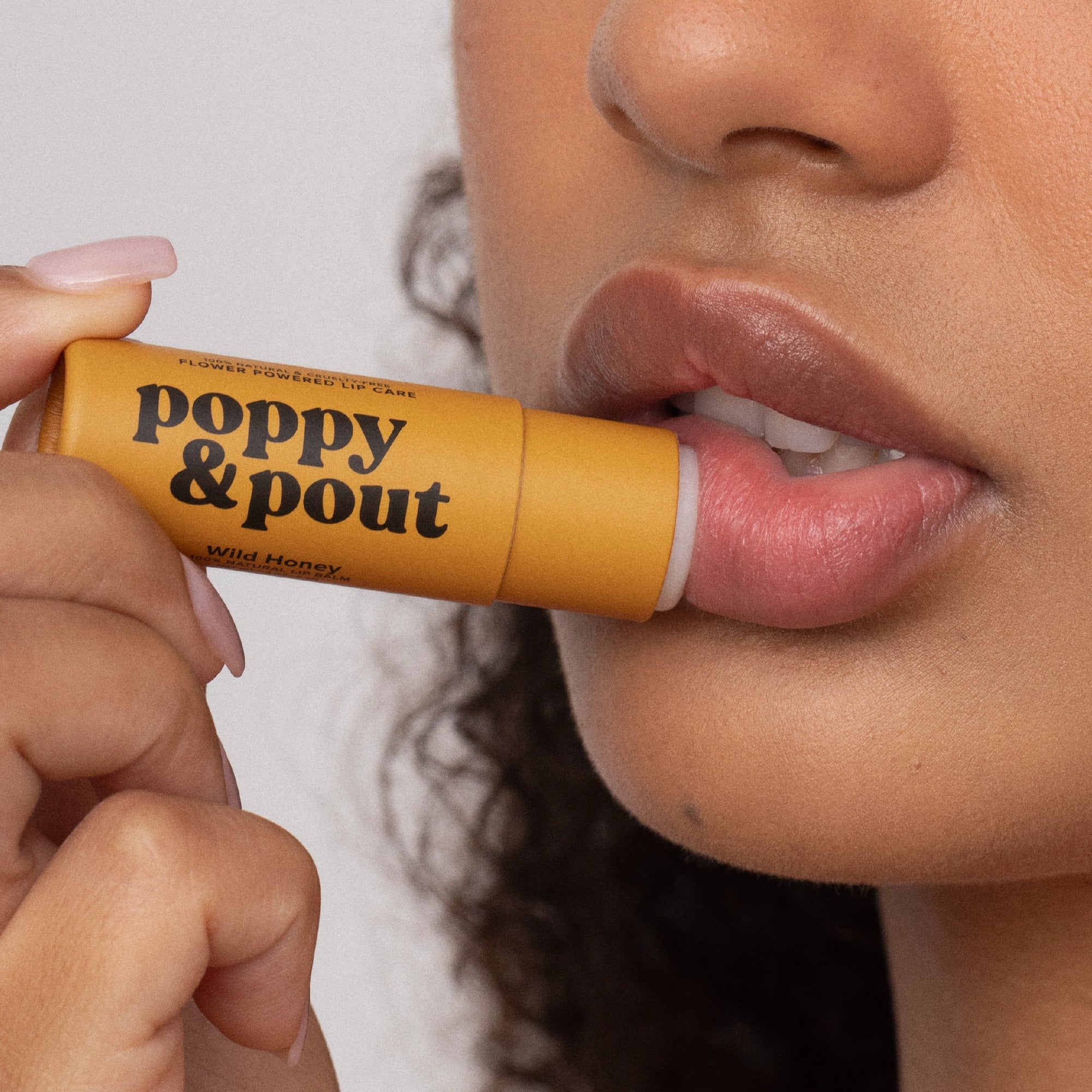 A person with curly hair applies a yellow tube of Faire Poppy & Pout Lip Balm to their lips, holding the eco-friendly packaging close to their mouth.