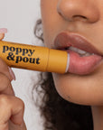 A person with curly hair applies a yellow tube of Faire Poppy & Pout Lip Balm to their lips, holding the eco-friendly packaging close to their mouth.
