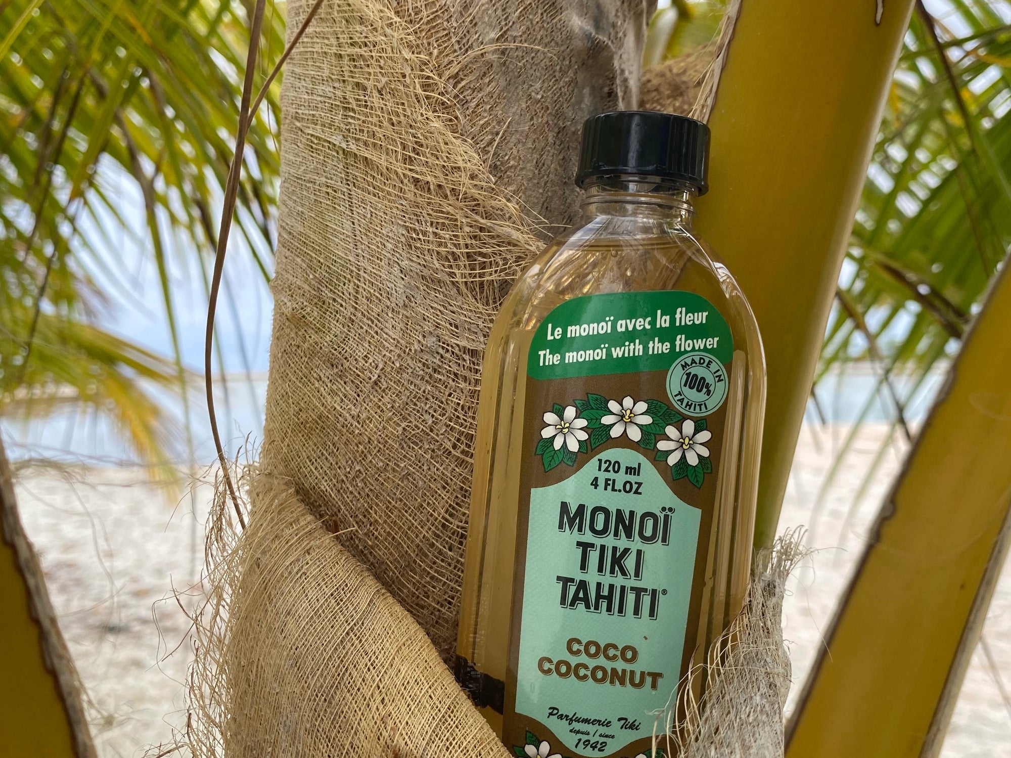 A bottle of Monoi Coco Coconut Oil by Faire leans against a palm tree trunk, wrapped in burlap, with lush green palms and sandy beach behind—showcasing the pure essence of Monoi Oil from Faire.