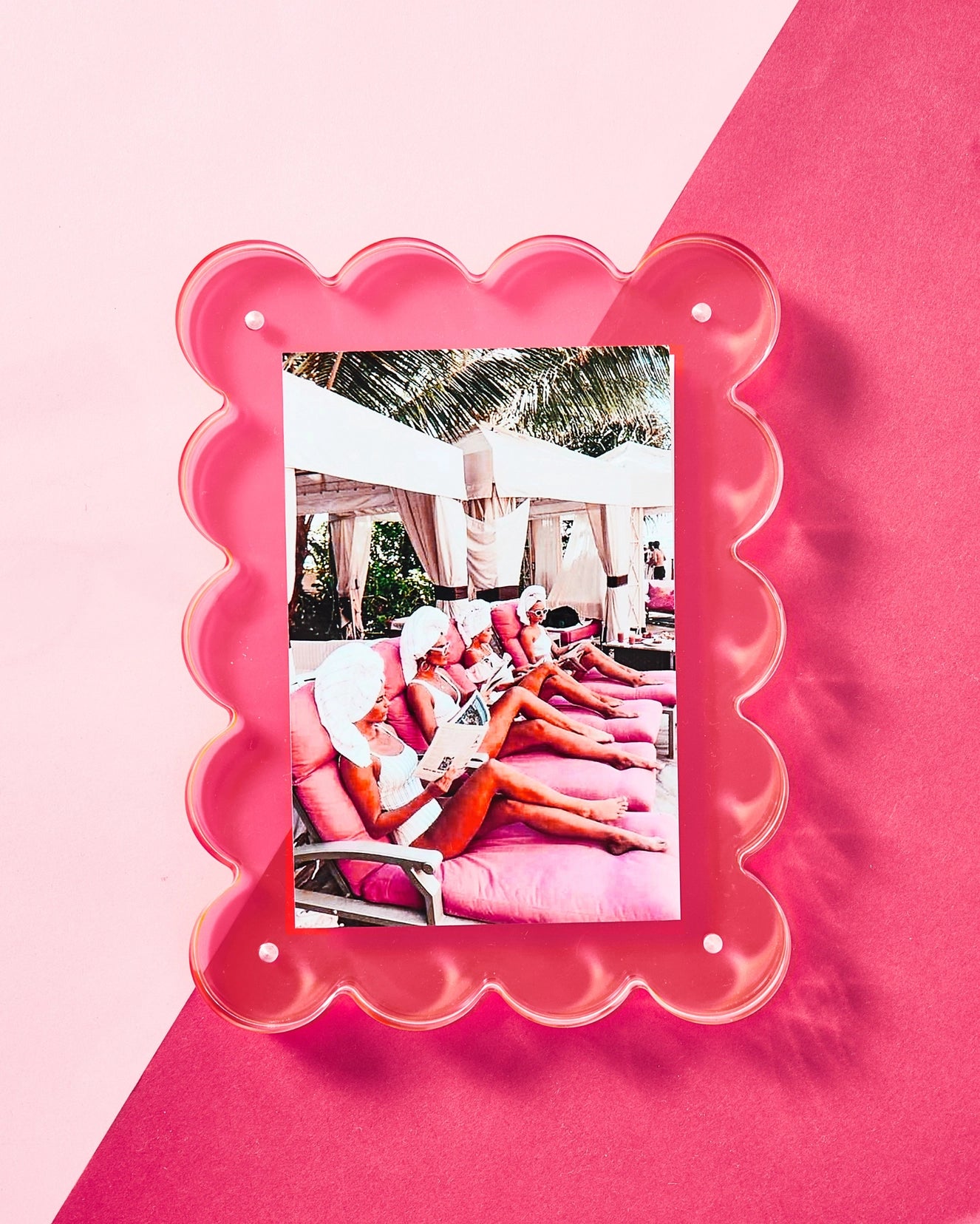 The Faire Acrylic Frame showcases a photo of people in pink swimsuits and white sun hats lounging on pink chairs. Set outdoors under cabanas amidst palm trees, the scene features a pink and light pink split background. The frame has decorative scalloped edges.