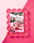 The Faire Acrylic Frame showcases a photo of people in pink swimsuits and white sun hats lounging on pink chairs. Set outdoors under cabanas amidst palm trees, the scene features a pink and light pink split background. The frame has decorative scalloped edges.